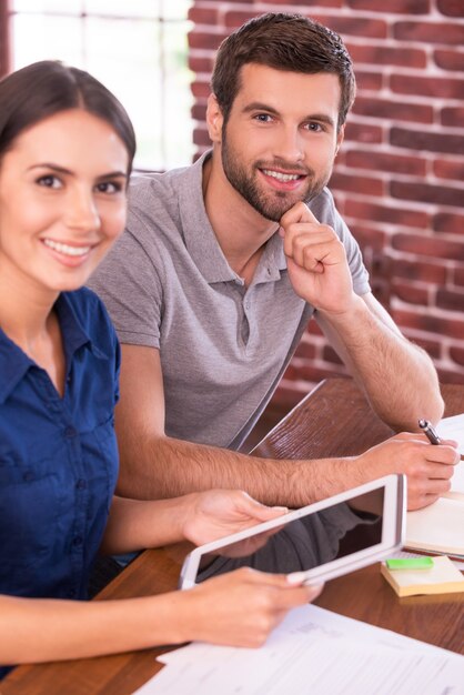 Young and successful. Top view image of cheerful young man and woman sitting at the working place and smiling while woman holding digital tablet