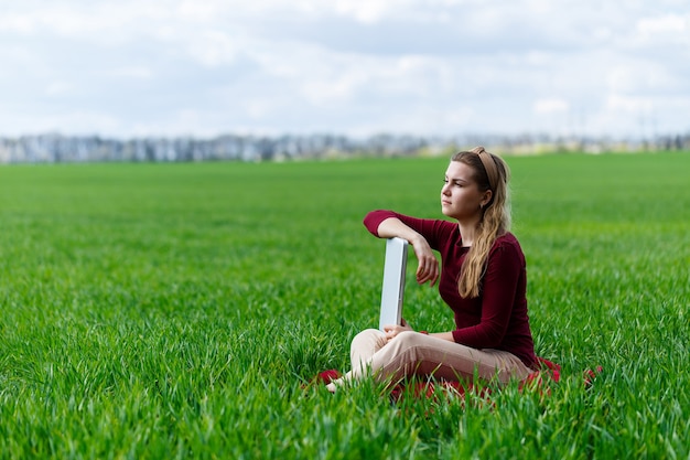 Young successful woman is sitting on green grass with a laptop in her hands. Rest after a good working day. Work on the nature. Student girl working in a secluded place. New business ideas