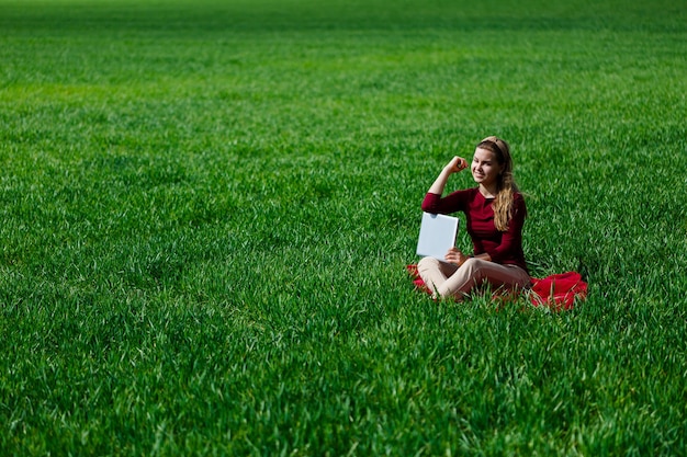 Photo young successful woman is sitting on green grass with a laptop in her hands. rest after a good working day. work on the nature. student girl working in a secluded place. new business ideas