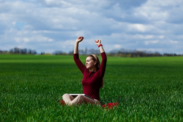 Young successful woman is sitting on green grass with a laptop in her hands. Rest after a good working day. Work on the nature. Student girl working in a secluded place. Workplace in nature