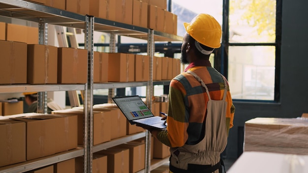 Young supervisor checking supplies on laptop, examining products inventory for merchandise on racks. Depot supervisor working on logistics in warehouse space, quality control cargo.