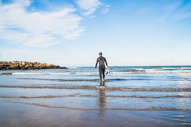 Young surfer entering into the water with his surfboard in a black surfing suit. Sport and water sport concept.