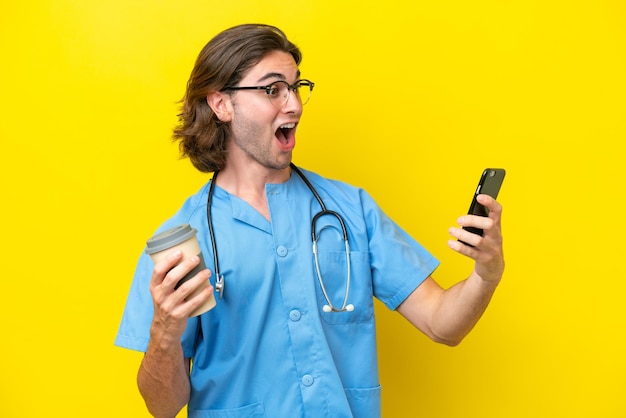 Young surgeon caucasian man isolated on yellow background holding coffee to take away and a mobile