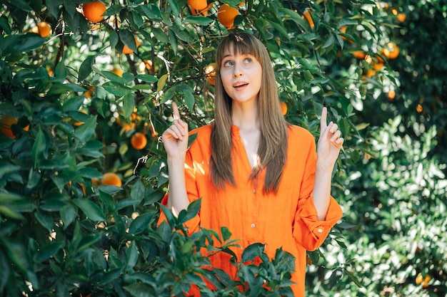 Young surprised girl in orange dress is looking and pointing up with forefinegrs in orange garden