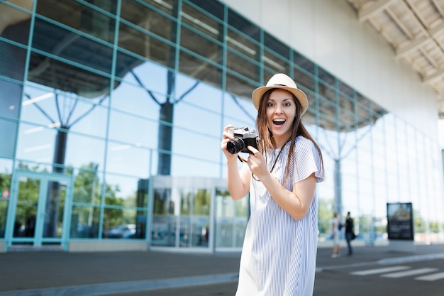 Young surprised joyful traveler tourist woman in hat holding retro vintage photo camera at international airport