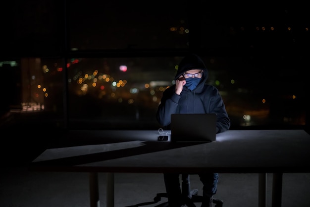 Young talented hacker using laptop computer while working in dark office with big city lights in the background at night