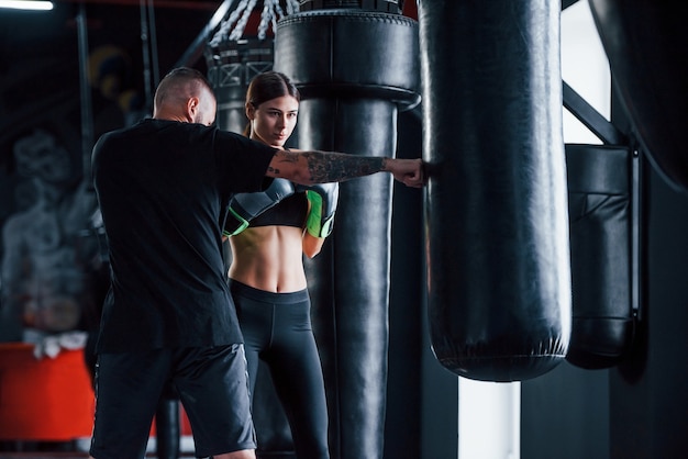 Young tattooed boxing coach teaches young woman in the gym.