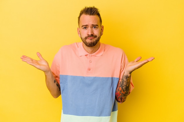 Photo young tattooed caucasian man isolated on yellow background doubting and shrugging shoulders in questioning gesture.