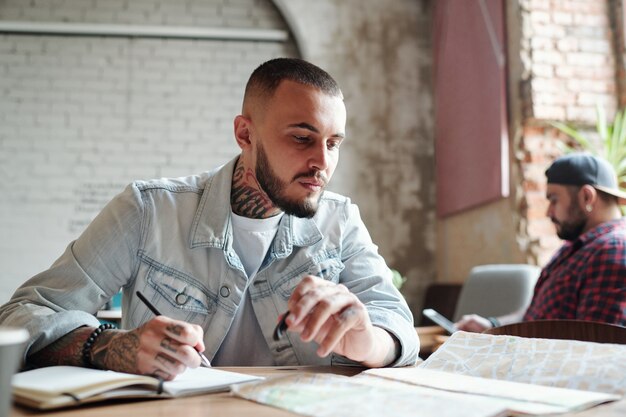 Young tattooed guy in denim jacket sitting at table in cafe and making notes while analyzing route using paper map