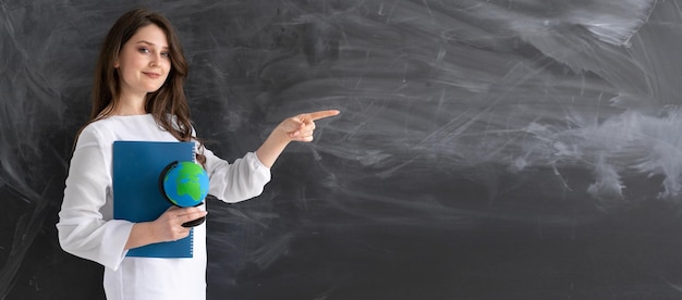 Young teacher or student millennial girl standing with a globe and a notebook in front of a black chalk board and pointing her finger at a blank space for text Banner School education concept