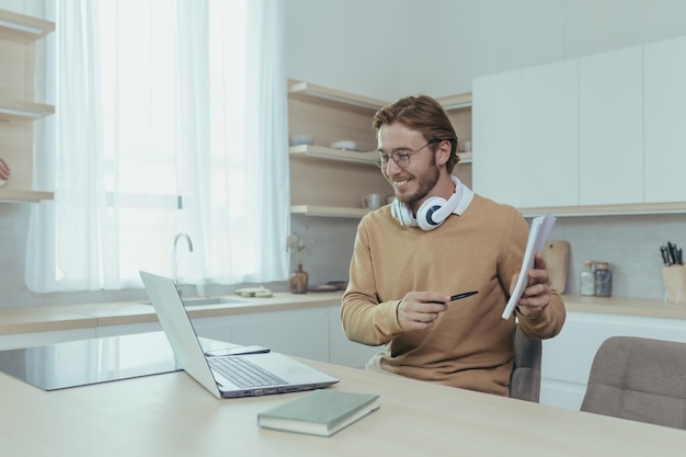 Young teacher teaches remotely from home man works in kitchen at home uses laptop for online call