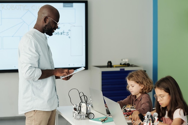 Young teacher with tablet standing in front of schoolchildren with laptops