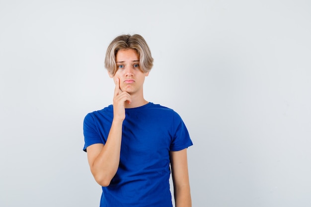 Young teen boy in blue t-shirt holding finger on cheek and looking dismal , front view.