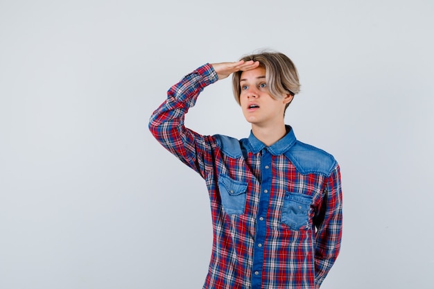 Young teen boy in checked shirt looking far away with hand over head and looking focused , front view.