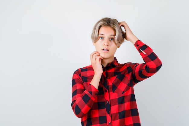 Young teen boy scratching head, touching his jaw in checked shirt and looking puzzled. front view.