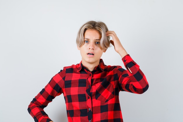 Young teen boy scratching his head while opening mouth in checked shirt and looking confused , front view.