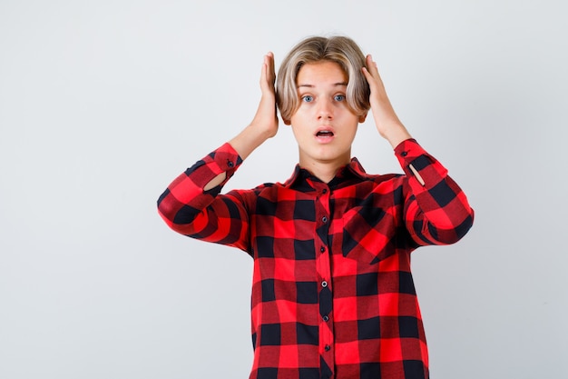 Young teen boy with hands near head in checked shirt and looking scared , front view.