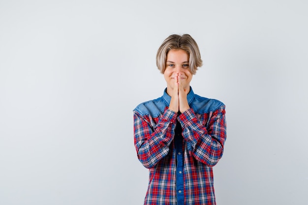 Young teen boy with hands in praying gesture in checked shirt and looking cheerful