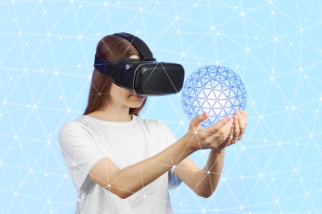 Young teen girl in a virtual reality helmet on a blue background is holds a three-dimensional sphere