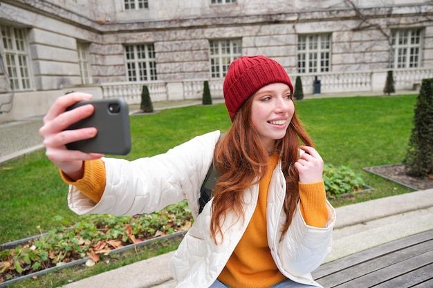 Young teen redhead girl sits on bench in park and takes selfie makes a photo of herself with smartph