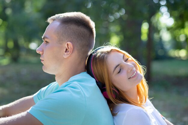 Young teenage couple together outdoors in summer park. Girl with red hair listening to music in earphones and boy is bored.