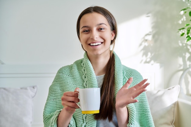 Young teenage female under knitted plaid warming up with hot tea in mug looking at camera