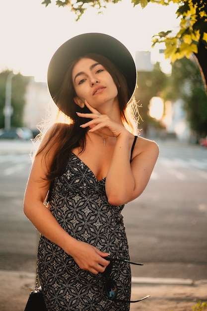 Young teenage girl in a white dress and hat. style of the big city. Sunset sky and rays of the sun. Natural beauty pretty woman in black fedora hipster hat, on warm summer evening sunset in big city