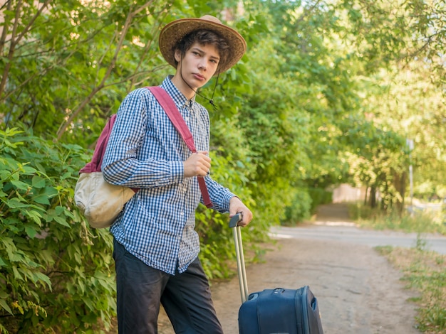 Young teenager boy in summer hat and casual clothes walking with suitcase and put on backpack 