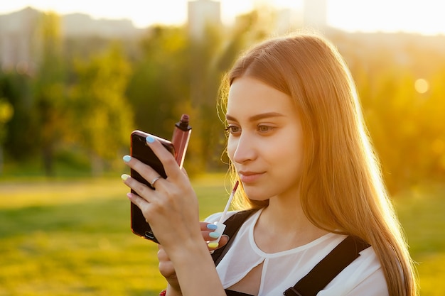 Young teenager girl hair fresh skin and a pretty face corrects make-up lipstick looking at the reflection in the phone in the park at sunset