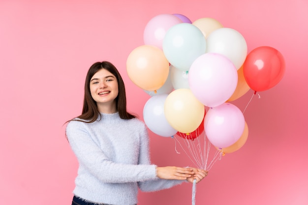 Young  teenager girl holding lots of balloons over pink wall applauding