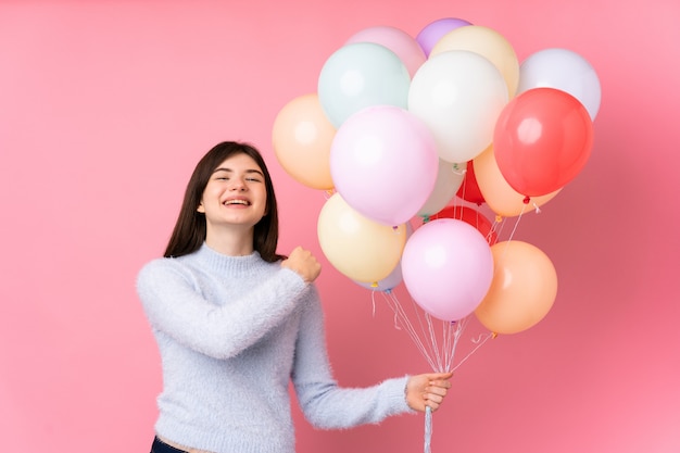 Young teenager girl holding lots of balloons over pink wall celebrating a victory