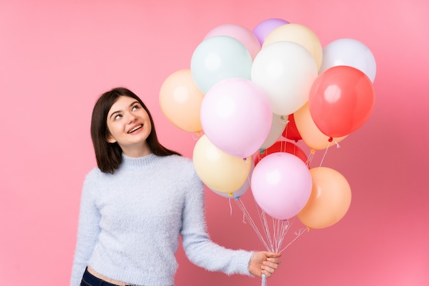 Young  teenager girl holding lots of balloons over pink wall laughing and looking up