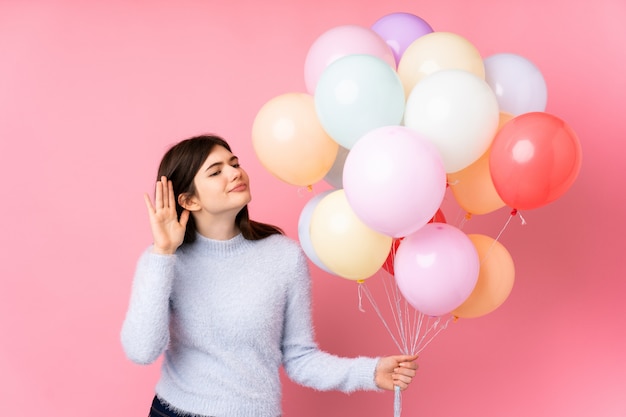 Young  teenager girl holding lots of balloons over pink wall listening something