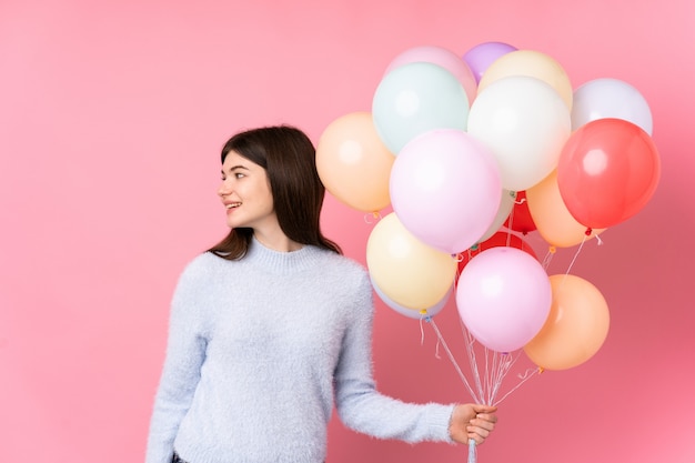 Young teenager girl holding lots of balloons over pink wall looking to the side