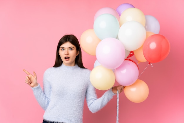 Young  teenager girl holding lots of balloons over pink wall surprised and pointing finger to the side