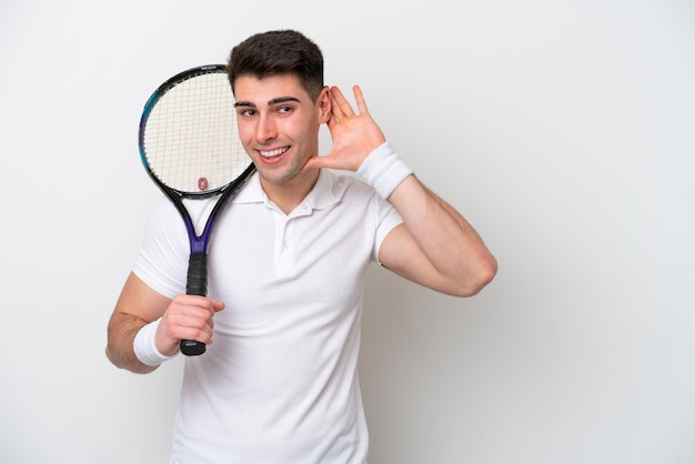 Young tennis player man isolated on white background listening to something by putting hand on the ear