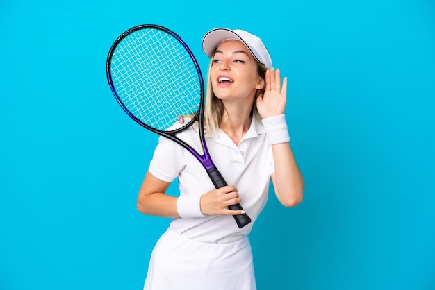 Young tennis player Romanian woman isolated on blue background listening to something by putting hand on the ear