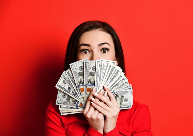 Young thinking pretty woman in red dress hiding behind bunch of money banknotes