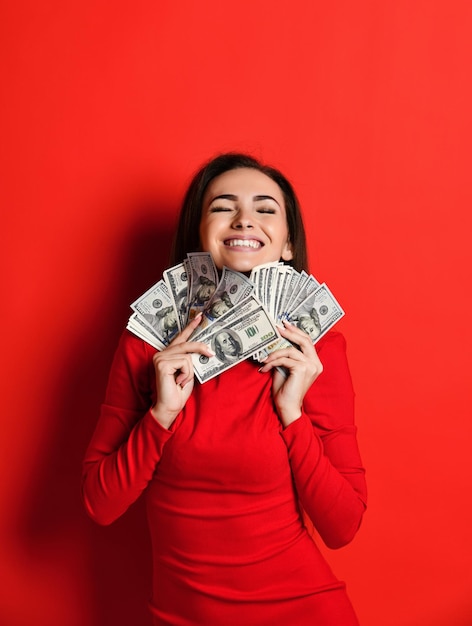 Young thinking pretty woman in red dress presses to herself a bunch of money banknotes