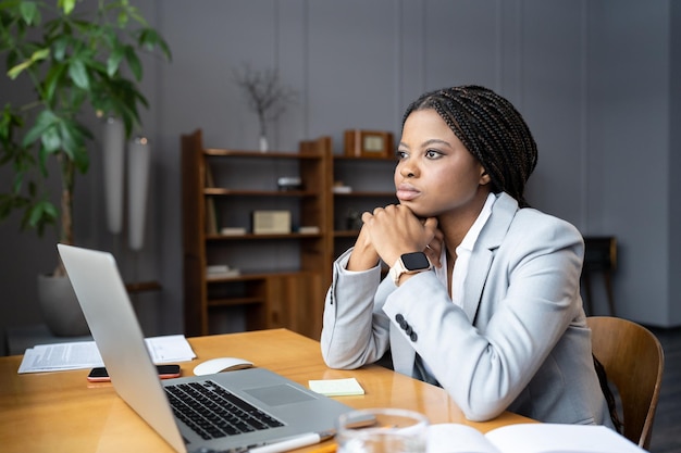 Young thoughtful african female employee looking out window at workplace and dreaming about vacation