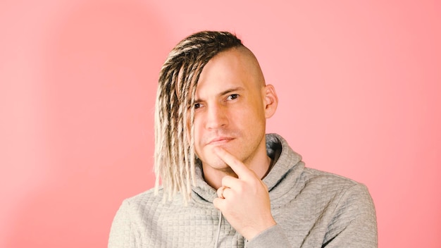 Young thoughtful handsome man with blonde dreadlocks on pink background Pensive guy with doubt glance taps his chin with finger