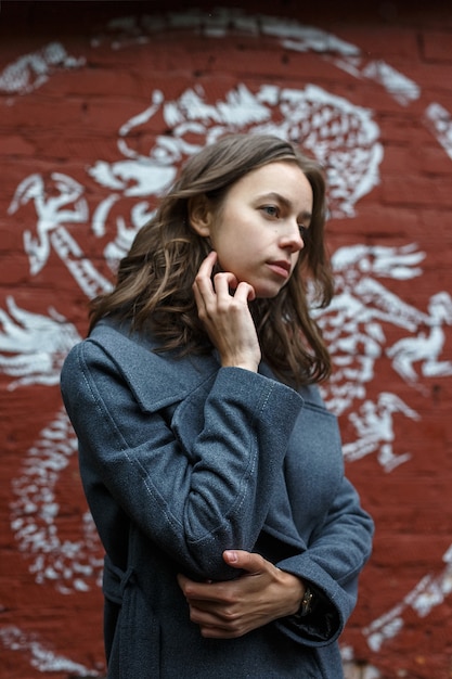 Young thoughtful pretty girl in gray coat and turtleneck poses in front of red brick wall with graffiti