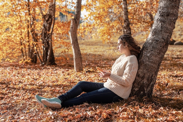 Young thoughtful woman sitting by the tree in autumn forest