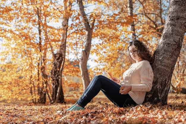 Young thoughtful woman sitting by the tree in autumn forest