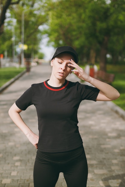 Young tired athletic beautiful brunette woman in black uniform and cap standing, resting and keeping hand near head after running, training in city park outdoors