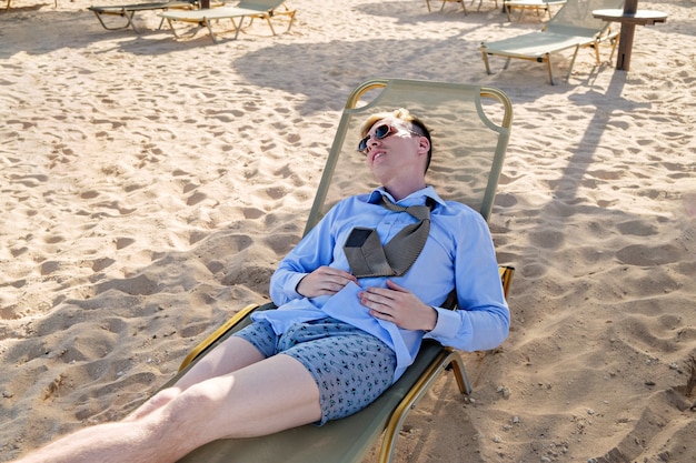 Young tired businessman lies on a chaise longue in a shirt with a tie and boxer shorts rest at sea r...