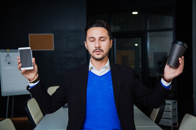 Young tired businessman meditating on table with mobile phone and cup of coffee in spacious office