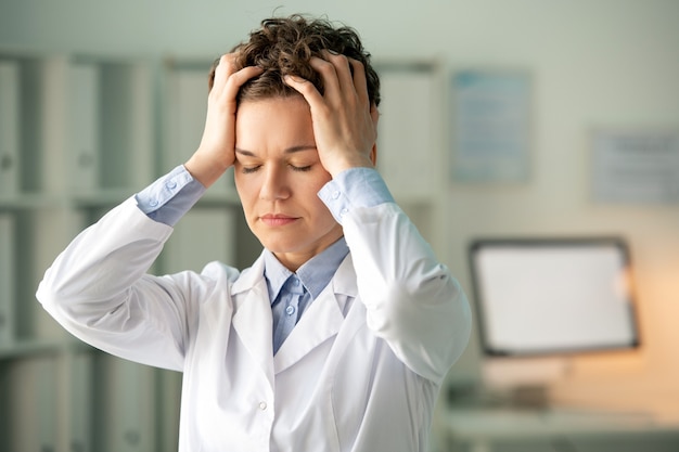 Young tired female chemist or pharmacist in whitecoat touching her head while standing against workplace in laboratory