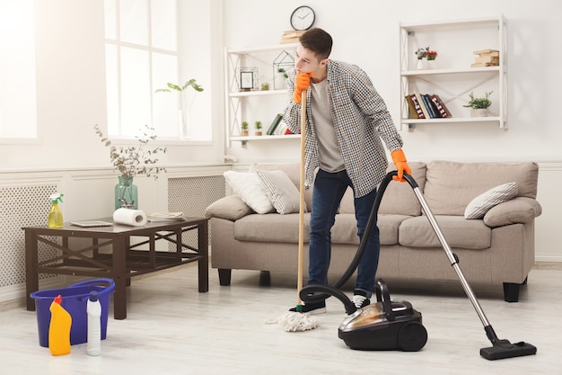 Young tired man cleaning home with mop and vacuum cleaner in living-room. Housekeeping and home cleaning concept