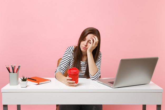 Young tired sleeping woman leaning on hand holding cup of coffee or tea while work and sit at office with pc laptop isolated on pastel pink background. Achievement business career concept. Copy space.